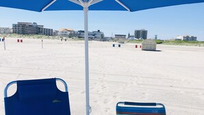 Ein Strand mit zwei blauen Liegestühlen unter einem blauen Sonnenschirm, einem Metalltisch und einem kleinen Gebäude im Hintergrund.