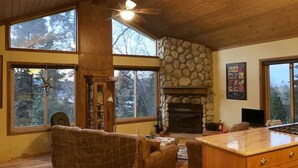 A spacious living room with a stone fireplace, large windows, and a ceiling fan. - Peaceful Cozy Cabin Amongst the Trees with View of Lake Arrowhead (Lake Arrowhead)