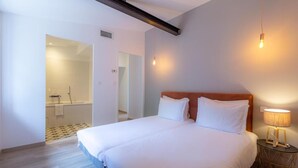 Quarto de hotel moderno com uma cama grande, uma mesa de cabeceira com abajur e um banheiro visível através de uma porta aberta.