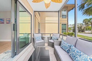 A modern balcony with wicker furniture and a sofa, large glass doors, and a view of a building and palm trees.