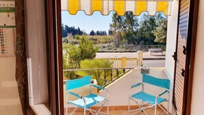 Een balkon met twee blauwe stoelen, een geel-wit gestreept zonnescherm en uitzicht op bomen en een hek.