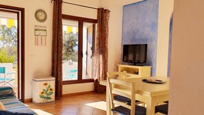 Een kamer met een bank, een eettafel met stoelen, een televisie, een kleine prullenbak en een raam met gordijnen.