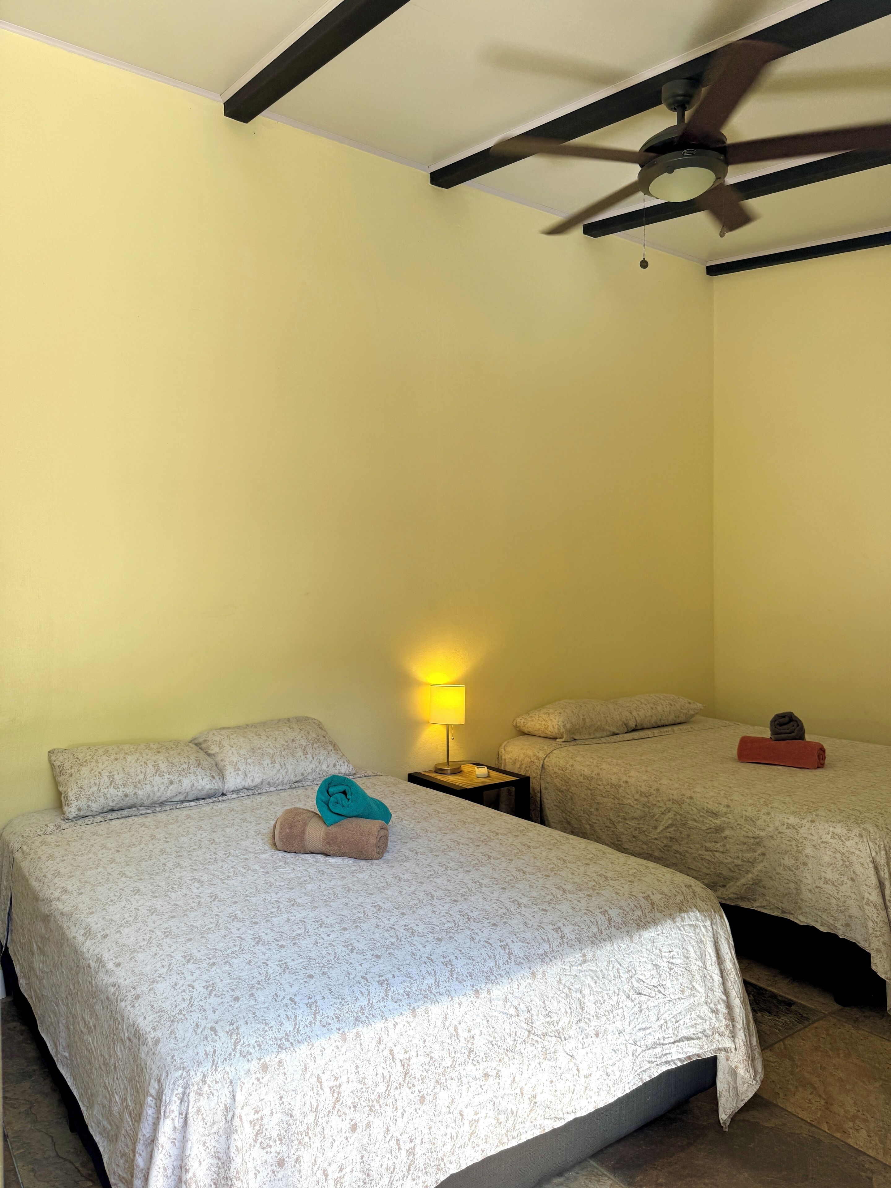 A hotel room with two beds, a ceiling fan, a nightstand with a lamp, and a red towel on the floor.