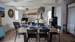 Ein Essbereich mit einem Tisch für sechs Personen, Blick auf den Strand durch ein großes Fenster und eine gemütliche Wohnarea mit einer Couch und einem Fernseher.