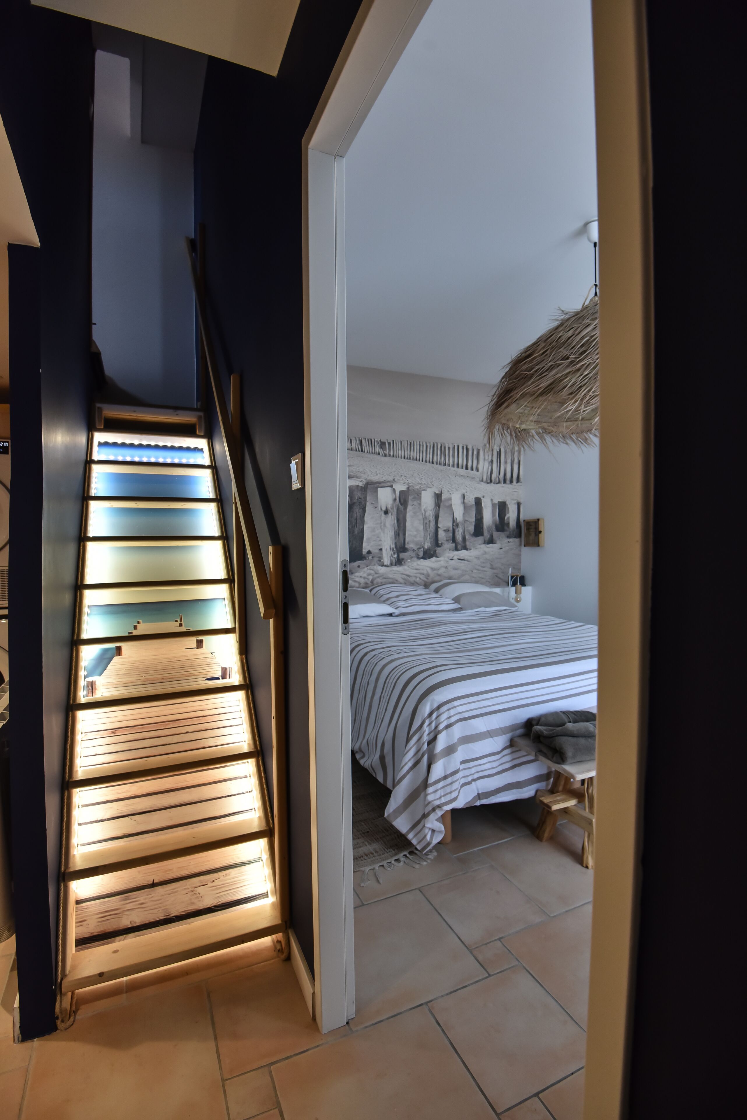 A bedroom with a bed, a nightstand, a wall-mounted artwork, and a staircase with illuminated steps.