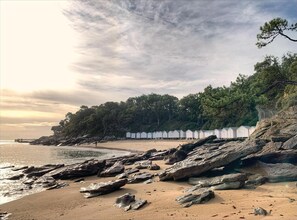 Une plage avec des cabanes de plage blanches, une côte rocheuse et des arbres en arrière-plan.