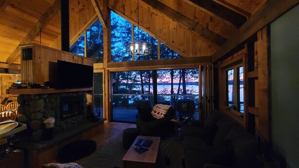 A cozy cabin interior with a stone fireplace, wooden walls, and large windows offering a view of the outdoors. - Cooper Bay Lakeside Escape – 322ft frontage, quiet, stunning west views! (Bracebridge)