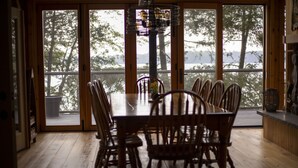 A dining area with a large wooden table and chairs, large windows with a view of trees and a body of water. - Cooper Bay Lakeside Escape – 322ft frontage, quiet, stunning west views! (Bracebridge)