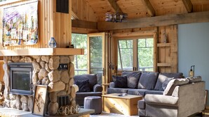 A cozy cabin interior with a stone fireplace, wooden walls, and a comfortable seating area with a couch and ottoman. - Cooper Bay Lakeside Escape – 322ft frontage, quiet, stunning west views! (Bracebridge)