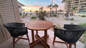 A wooden table with two black wicker chairs on a tiled patio overlooking a beach with palm trees and a building in the background. - Beachfront Retreat with Convenient Access to Shops, Dining, and More (San José del Cabo)