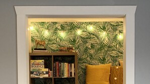 A framed view of a cozy reading nook with a bookshelf, a plush sofa, and a tropical leaf-patterned wallpaper. - A Charming Home Minutes to Downtown, Zoo, & Airport with Hidden Reading Nook! (Des Moines)