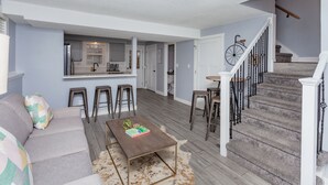 A modern living room with a sofa, coffee table, and bar stools. A staircase with a railing leads to a second floor. - A Charming Home Minutes to Downtown, Zoo, & Airport with Hidden Reading Nook! (Des Moines)