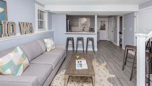 A living room with a sofa, coffee table, and a rug. There is a kitchen in the background with a window and a refrigerator. - A Charming Home Minutes to Downtown, Zoo, & Airport with Hidden Reading Nook! (Des Moines)