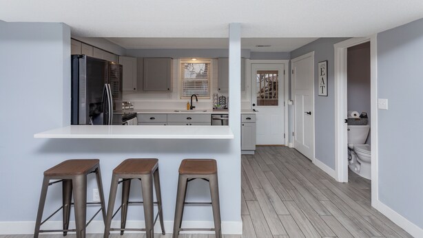 A modern kitchen with a central island, bar stools, and a view into the adjacent bathroom. - A Charming Home Minutes to Downtown, Zoo, & Airport with Hidden Reading Nook! (Des Moines)