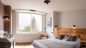 A bedroom with a large window, a bed with patterned bedding, a wooden headboard, and a small bedside table.