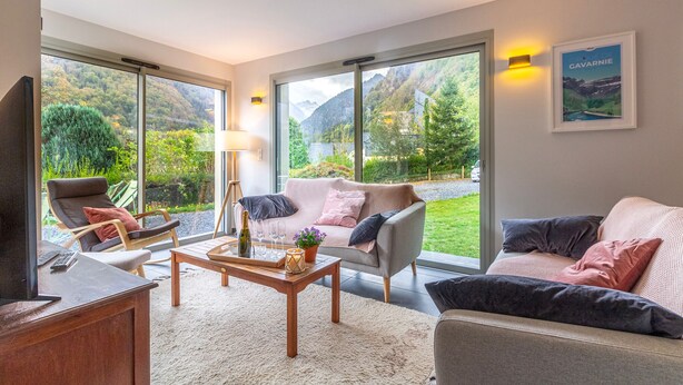 A modern living room with a sofa, armchairs, a coffee table, and a TV. Large windows offer a view of a mountainous landscape.