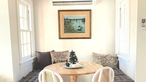 A cozy dining area with a round wooden table, two white wicker chairs, and a bench with cushions. An air conditioning unit is mounted on the wall above the table. - Renovated cabin / beach access / 1+ acre of woods (South Haven)