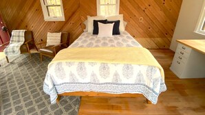 A bedroom with a wooden sloped ceiling, a bed with patterned bedding, a chair, a desk, and a window. - Renovated cabin / beach access / 1+ acre of woods (South Haven)