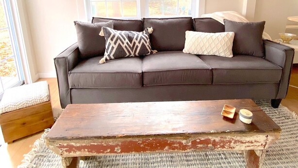 A living room with a dark brown sofa, two windows, a wooden coffee table, and a woven rug. - Renovated cabin / beach access / 1+ acre of woods (South Haven)