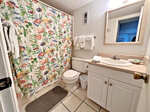 A bathroom with a shower curtain featuring a floral pattern, a toilet, a sink with a mirror above it, and a small trash bin. - 🌴You CAN afford Paradise! Life is better at the beach. 🌊Call today! (Gulf Shores)