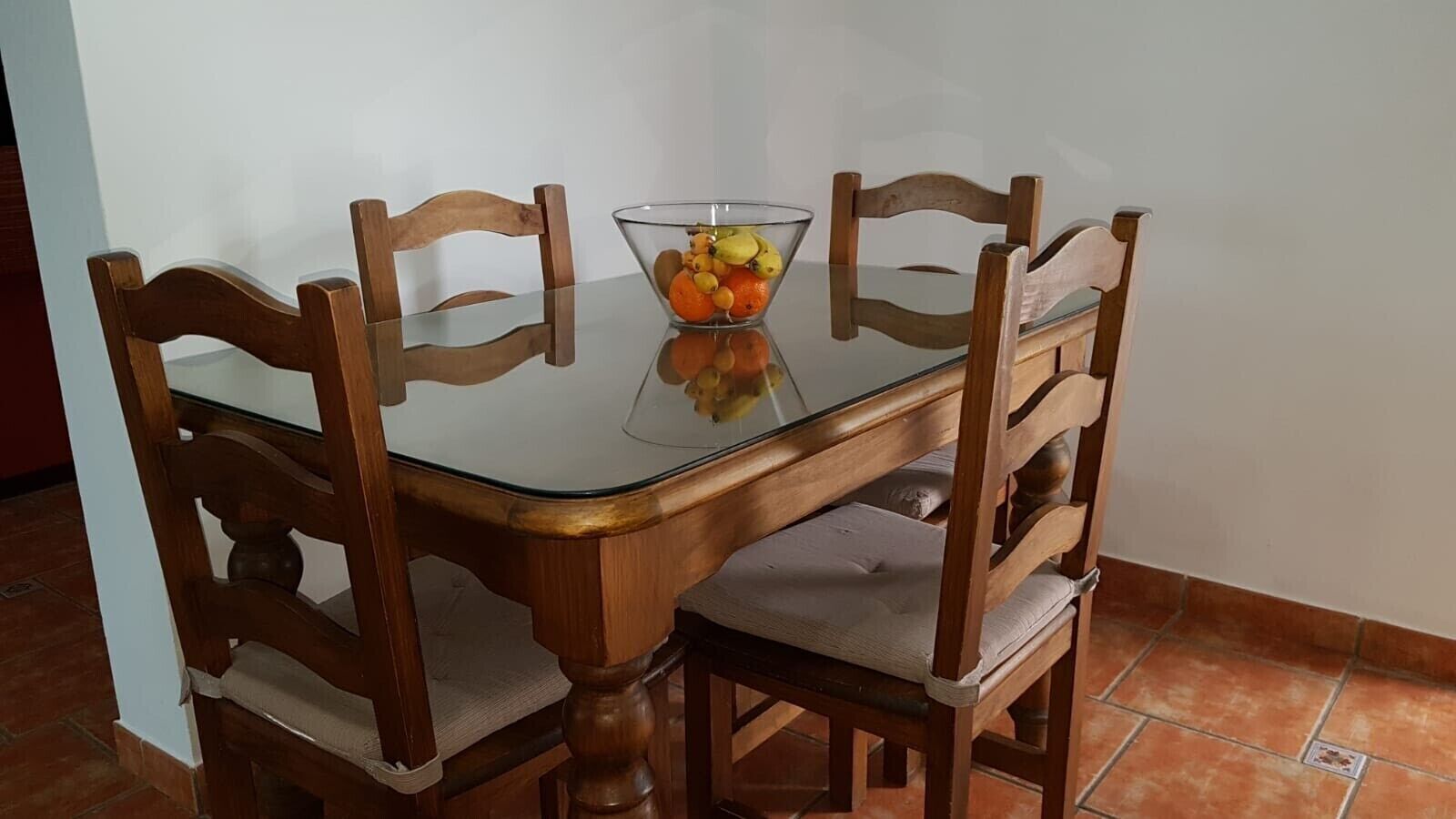 A dining table with four wooden chairs, a glass top, and a bowl of fruit on the table.