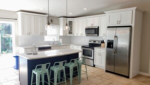A modern kitchen with white cabinets, a stainless steel refrigerator, a microwave, and a stove. There are three green bar stools around the island. - Mystical - Relaxing Heated Pool and Stylish Comfortable Home, Pet Friendly! (St. Augustine)