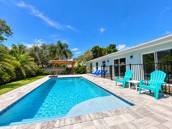 A rectangular swimming pool with a wooden deck and lounge chairs, surrounded by a white building with large windows and a pergola.