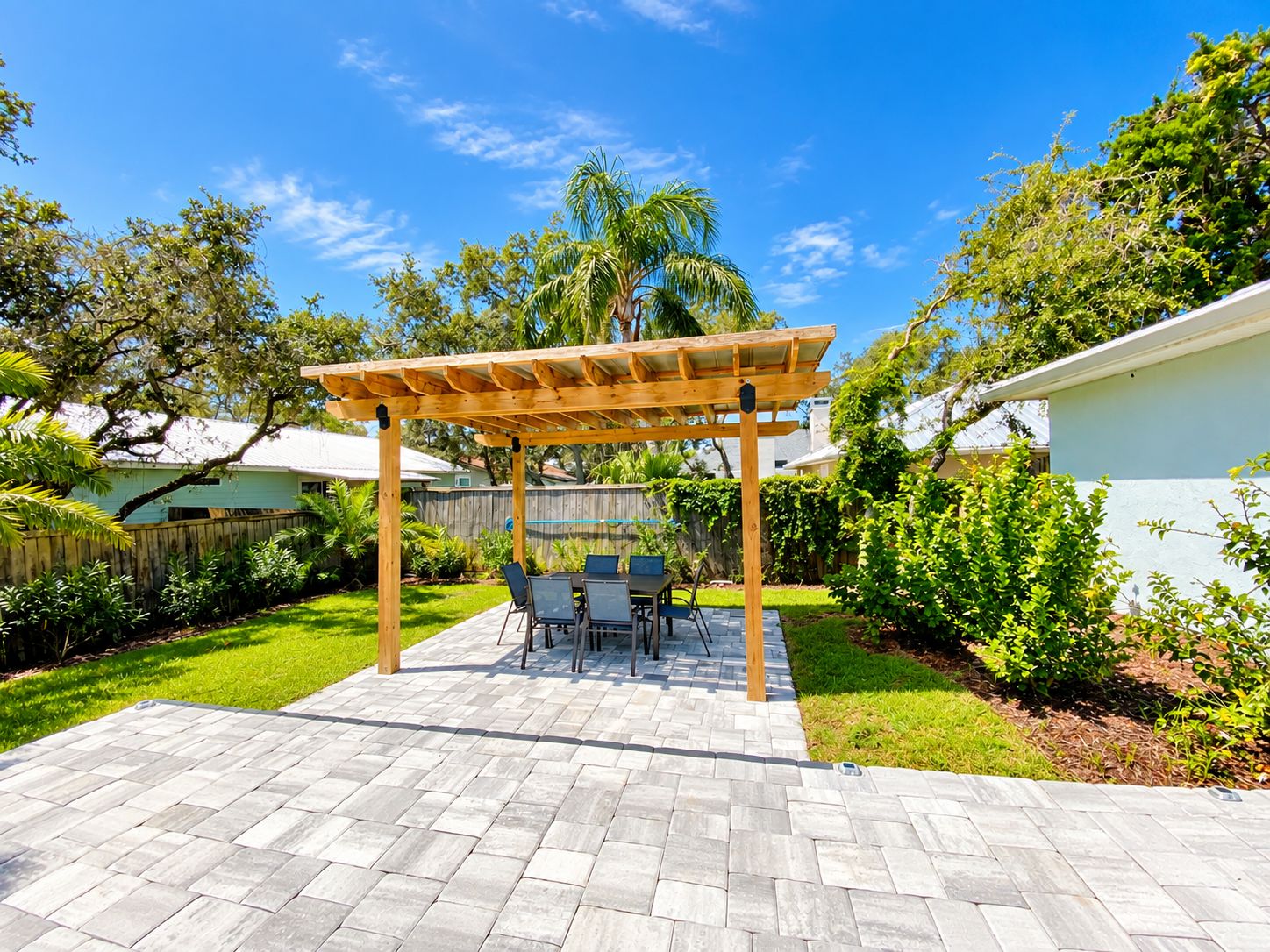 A covered patio area with a table and chairs, surrounded by a wooden pergola, a paved patio, and a grassy lawn.