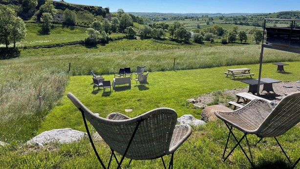 Ein grasbewachsenes Freigelände mit Korbstühlen und Picknicktischen, mit Blick auf eine malerische Landschaft aus Hügeln und Bäumen.