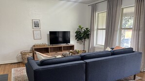 Un salon avec un canapé bleu foncé, un téléviseur à écran plat posé sur un meuble en bois, une plante en pot et une fenêtre avec des rideaux.