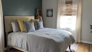 Une chambre à coucher avec un lit, une tête de lit, une échelle en bois, un tableau encadré et une fenêtre avec des rideaux.