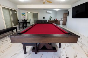 Une table de billard dans une pièce dotée d’un ventilateur de plafond, d’un téléviseur et d’un coin cuisine en arrière-plan.