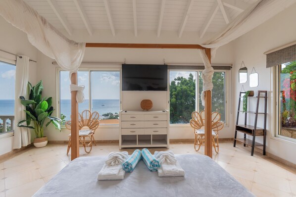 A bedroom with a large bed, two wicker chairs, a dresser, and a view of the sea.