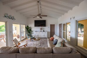 A spacious living room with a ceiling fan, a flat-screen TV mounted on the wall, a sofa, a coffee table, and a dining area with a table and chairs.
