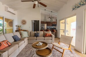 A spacious living room with a sofa, coffee table, and a chair. There is a kitchen in the background with a refrigerator and microwave.