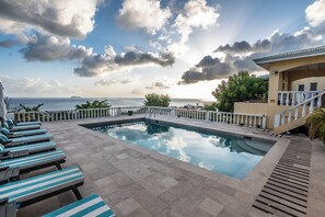 A swimming pool with lounge chairs, a view of the sea, and a building with a balcony.