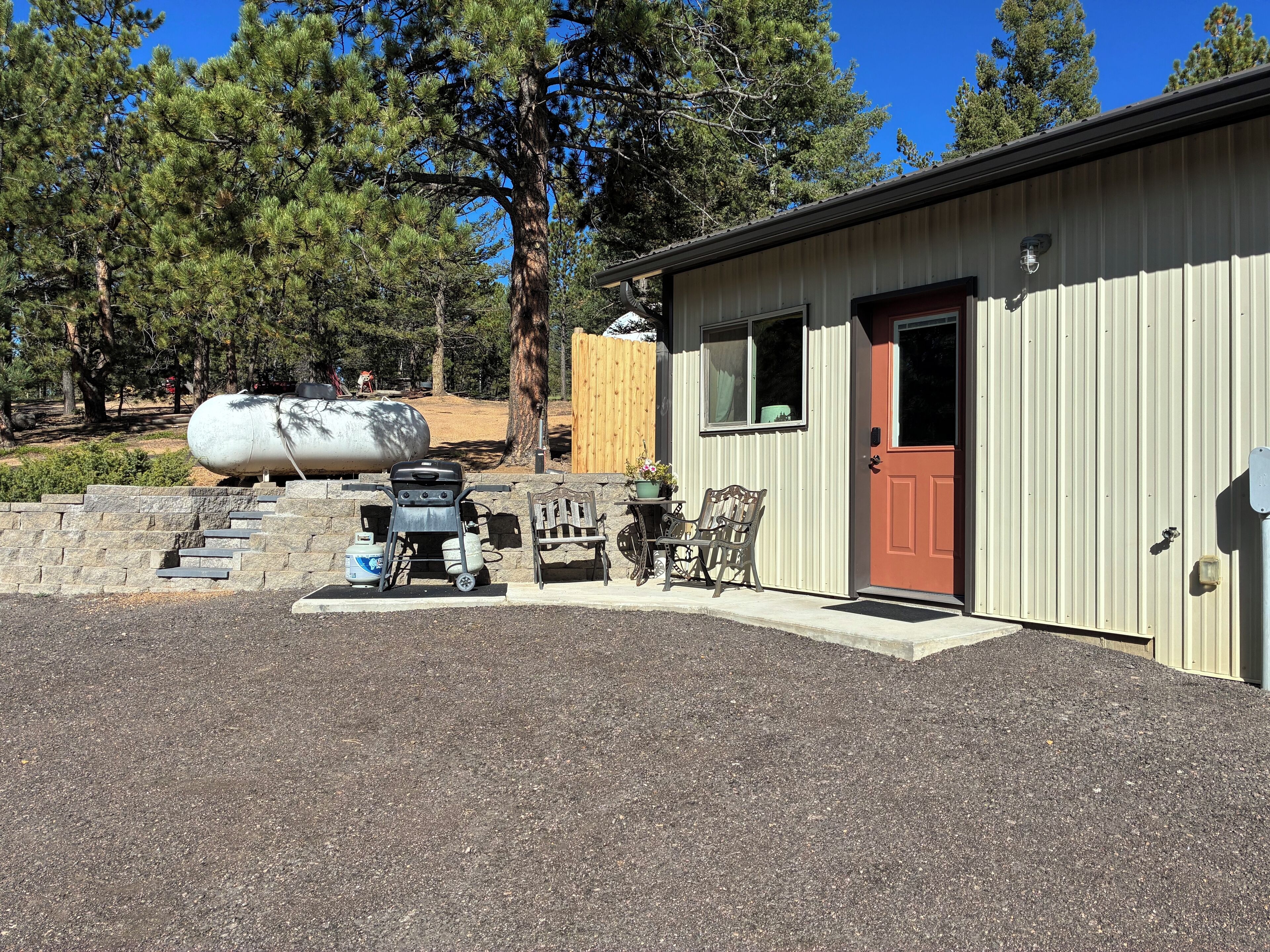 A building with a red door, a small patio with a grill and chairs, and a large propane tank in the background.