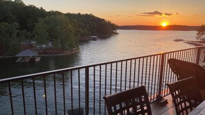 Une vue sur le coucher de soleil depuis un balcon doté d’une balustrade, donnant sur une rivière paisible où l’on aperçoit des bateaux et un quai.