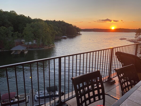A sunset view from a balcony with a railing, overlooking a calm river with boats and a dock.