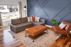 Relax in this stylish living room with a sofa, coffee table, and armchair, surrounded by a geometric-patterned rug and a large window offering a view of the outdoors.