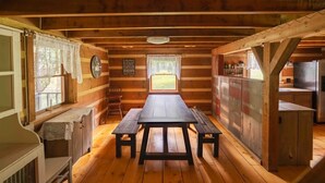A wooden cabin interior with a central table, benches, a window with curtains, a clock, and a kitchen area. - My Old Kentucky Home!  Restful Log cabin retreat.. (Madisonville)