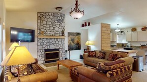 A living room with a stone fireplace, a television, a sofa set, and a kitchen area. - On Golf Course | Pet Friendly | Nama-Stay (Angel Fire)