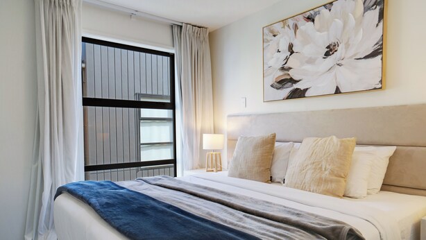 A hotel room with a large bed, a window with curtains, a wall-mounted painting, and a bedside lamp. - Brand New House by Botany - Parking (Auckland)
