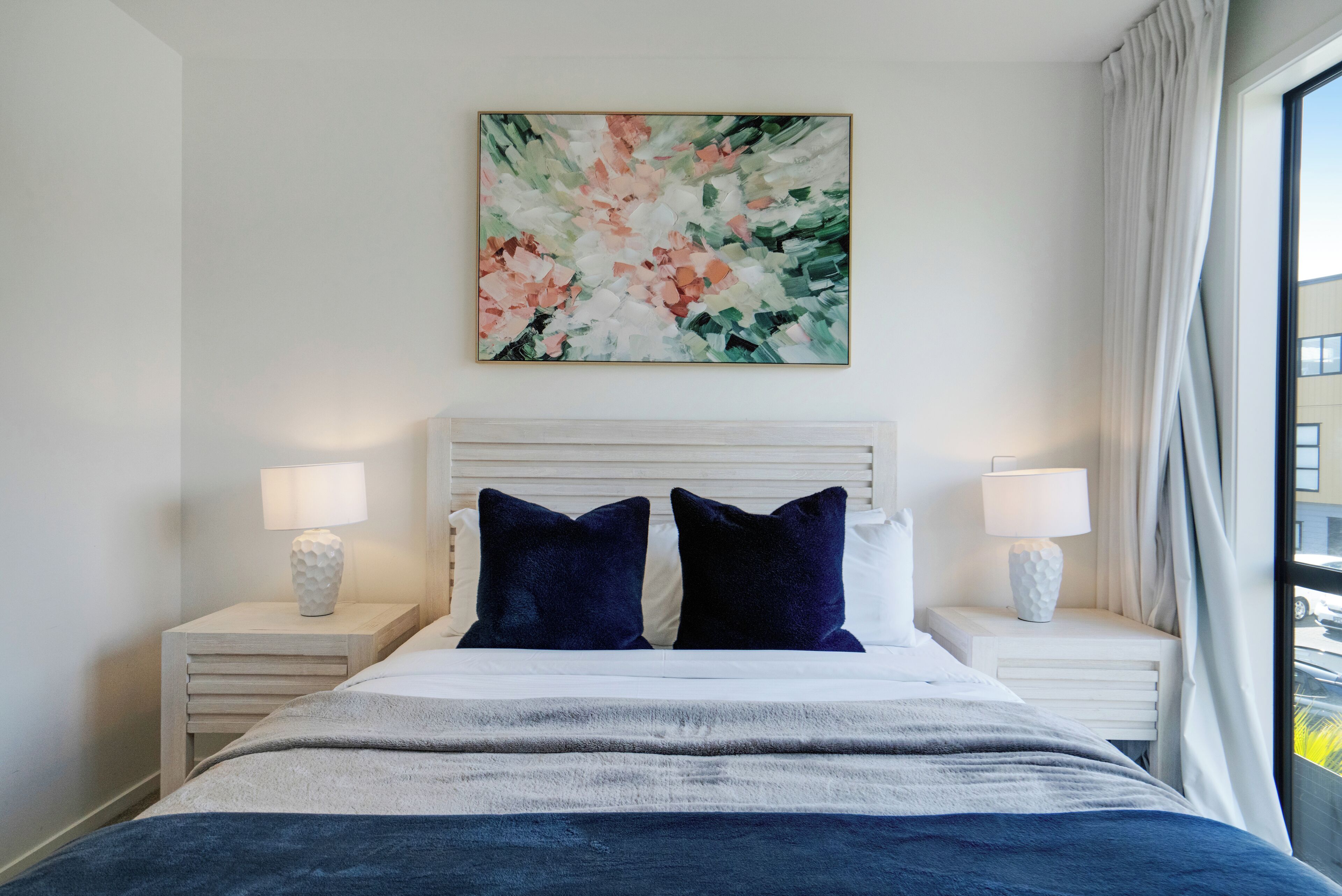 Un dormitorio con cabecera blanca, dos mesitas de noche con lámparas, una gran obra de arte floral en la pared y vistas al exterior a través de una ventana.