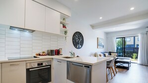 A modern kitchen with white cabinetry, a dishwasher, and an oven. There is a dining area with a table and chairs, and a view of the outside through large windows. - Brand New House by Botany - Parking (Auckland)
