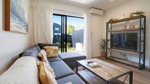 A modern living room with a sofa, a coffee table, and a TV on a wooden stand. - Brand New House by Botany - Parking (Auckland)