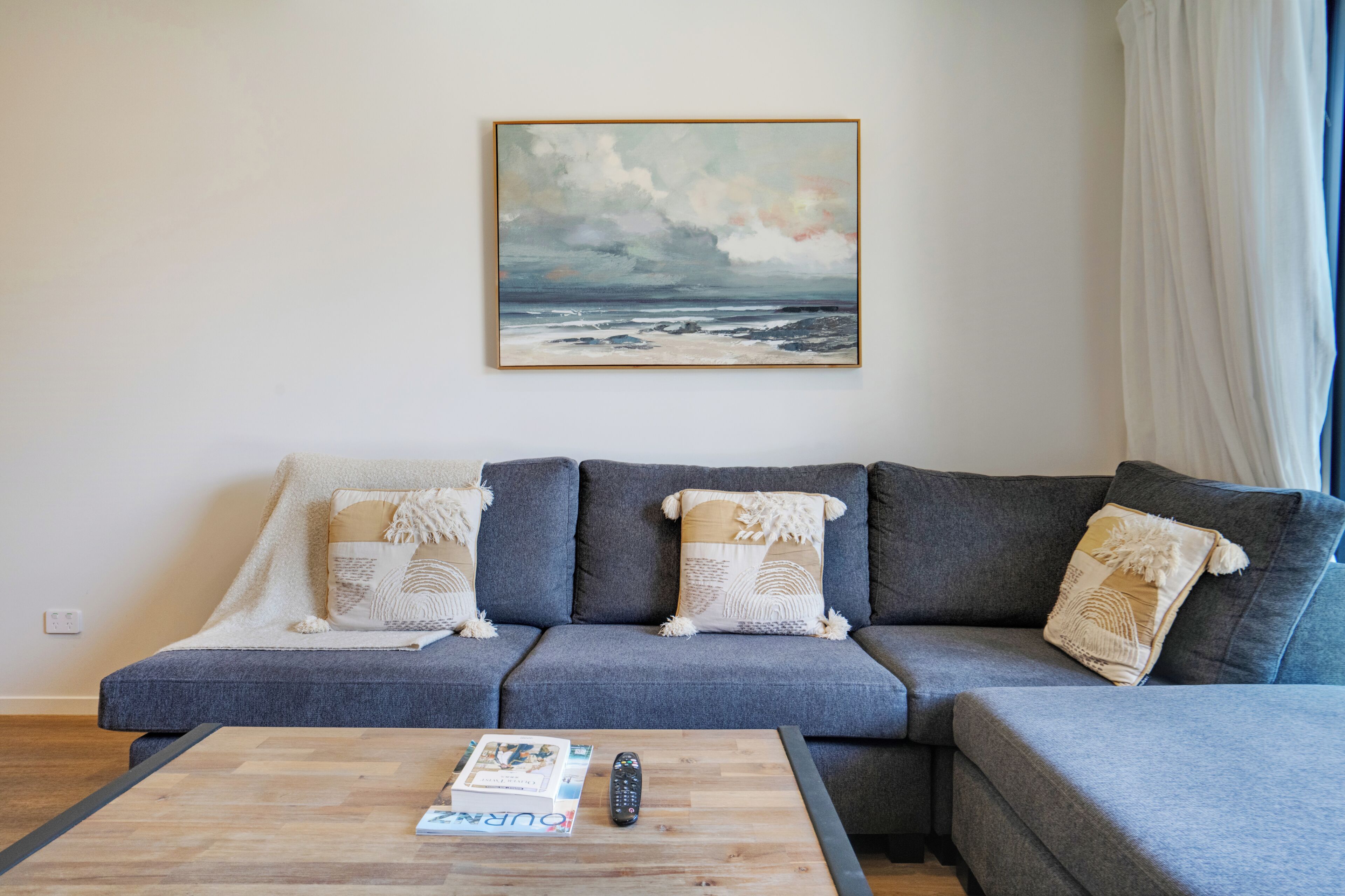 A living room with a grey sofa, a coffee table with a book and remote, and a painting of a cloudy sky.