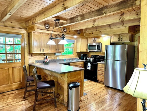 A wooden cabin-style kitchen with a central island, stainless steel appliances, and a view of the outdoors through large windows.