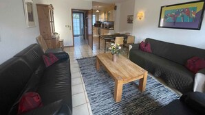 Una sala de estar con un sofá negro, una mesa de centro de madera y un sofá gris. Se ve una cocina al fondo con una mesa de comedor y sillas.