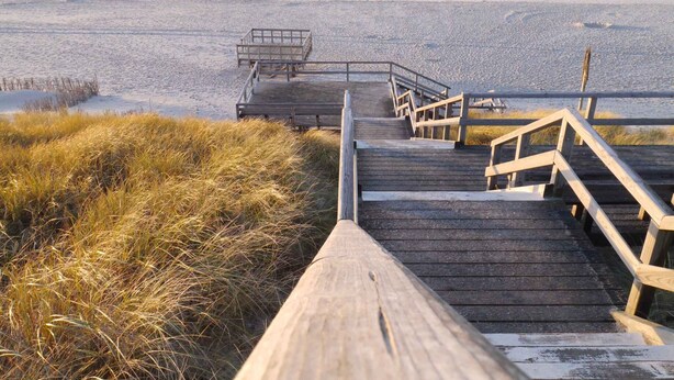 Un paseo marítimo de madera que conduce a una playa, con barandilla de madera y una pequeña estructura de madera.